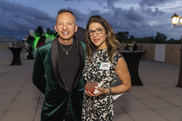 A man in a dark green velvet jacket stands next to a woman in a black-and-white patterned dress holding a drink. They attend the 2025 Men of Influence and Women of Influence event outdoors under a cloudy sky with decorative lights. Lifestyle