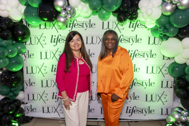 Two women of influence pose and smile in front of a "Lifestyle LUXE" backdrop, surrounded by green, black, and white balloon decorations. One wears a pink top and white pants; the other stands out in a bright orange outfit. Lifestyle