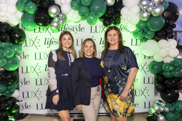 Three women, representing Women of Influence 2025, pose and smile in front of a "Luxe Lifestyle" backdrop, surrounded by green, black, white, and silver balloons arranged in an elegant arch. Lifestyle