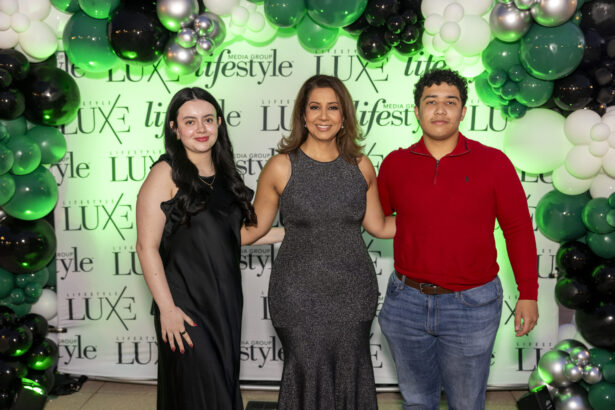 Three people, including a possible 2025 Women of Influence honoree, pose and smile in front of a Luxe Lifestyle Media Group backdrop adorned with black, white, and green balloons. The woman in the center wears a sleeveless dress; her companions wear black and red. Lifestyle