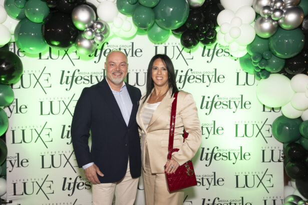 A smiling man in a navy suit and a woman in a beige suit with a red handbag stand together at the 2025 Women of Influence event, posed before a green, black, and white balloon arch and a backdrop with "LUXE" and "Lifestyle" logos. Lifestyle