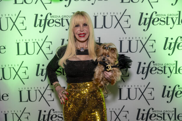A woman with long blonde hair, wearing a black top and shiny gold pants, smiles while holding a small dog in a black outfit, standing in front of a “Lifestyle Luxe” media group step-and-repeat backdrop at the 2025 Women of Influence event. Lifestyle