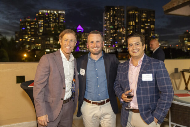 Three men of influence in business attire smile for a photo at an outdoor 2025 evening event, city skyline with lit-up buildings behind them; one holds a glass of wine and they all wear name tags. Lifestyle