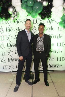 Two men in suits stand smiling in front of a green and white balloon arch and a backdrop with "LUXE" and "lifestyle" logos at the 2025 Men of Influence indoor event. Lifestyle