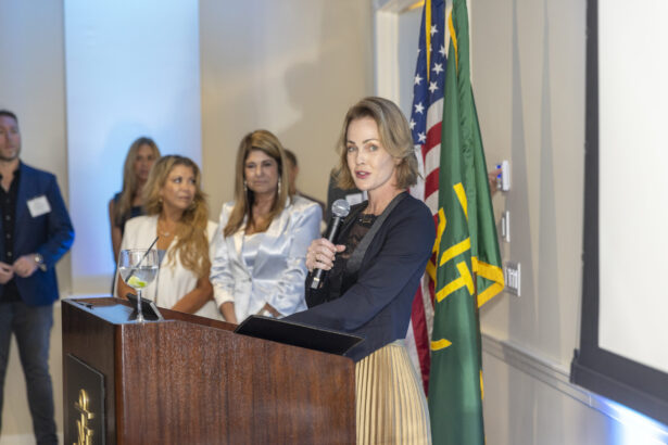 A woman speaks at a podium with a microphone, standing beside green and American flags, while several people listen in the background at the 2025 Women of Influence indoor event. Lifestyle