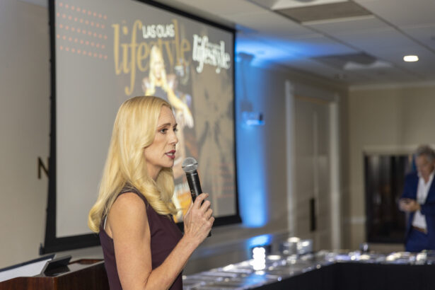 A woman with long blonde hair speaks into a microphone at an indoor 2025 event, standing near a screen displaying the word "lifestyle." The well-lit room has tables and another person blurred in the background. Lifestyle