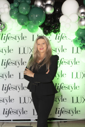 A woman with long blonde hair in a black suit stands smiling with arms crossed in front of an "Influential People 2025" media backdrop and green, white, and black balloons. Lifestyle
