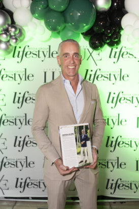 A man in a beige suit stands smiling, holding a magazine featuring his photo. Behind him, "lifestyle" is displayed on a backdrop with green, white, and black balloons—celebrating influential people 2025. Lifestyle