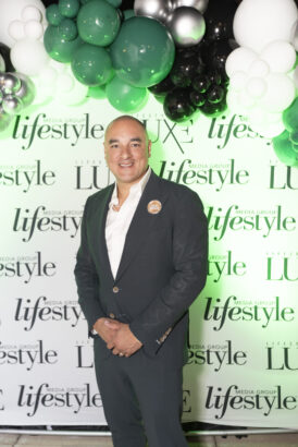 A man in a dark suit stands smiling in front of a "Lifestyle" and "Luxe" media group step-and-repeat backdrop, adorned with green, black, and white balloons at the 2025 Men of Influence event. Lifestyle