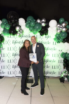 Two people stand smiling and holding a magazine in front of a backdrop with "LUXE" and "lifestyle" logos, decorated with black, white, and green balloons, celebrating the 2025 Men of Influence. Lifestyle