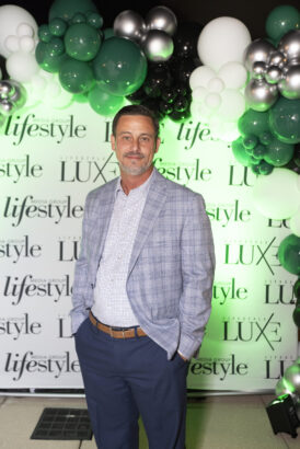 A man in a gray plaid blazer and blue pants stands before a "lifestyle" and "LUX" step-and-repeat banner at the 2025 Men of Influence event, with green, white, black, and silver balloons arranged in the background. Lifestyle