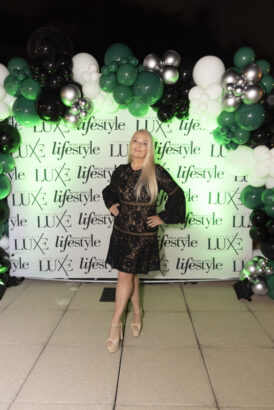 A woman in a black lace dress poses in front of a step-and-repeat banner with “LUXE” and “lifestyle” logos, surrounded by green, black, and white balloons at the 2025 Women of Influence event. Lifestyle