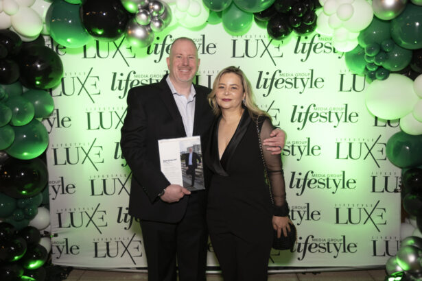 A man and a woman in formal attire stand smiling in front of a green, black, and white balloon arch with a "LUXE lifestyle" media backdrop at the Influential People 2025 event. The man holds a magazine or booklet. Lifestyle