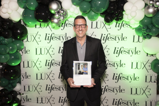 A smiling man in a suit holds an award plaque while standing before a step-and-repeat backdrop with "LUXE" and "lifestyle" logos, surrounded by white, black, and green balloons at the Men of Influence 2025 Influencers event. Lifestyle