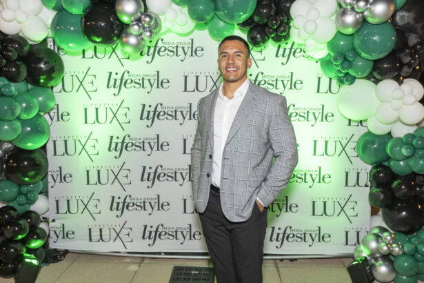 A man in a gray checkered blazer and white shirt, smiling, poses before a "LUX lifestyle" backdrop and a green, black, and silver balloon arch at the Influential People 2025 event. Lifestyle