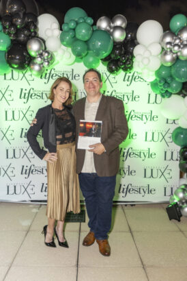 A woman and a man, both smiling, stand in front of a "Lifestyle" and "LUXE" step-and-repeat banner adorned with green, white, and silver balloons. The man holds a magazine, celebrating the 2025 Women of Influence and Influential People 2025. Lifestyle