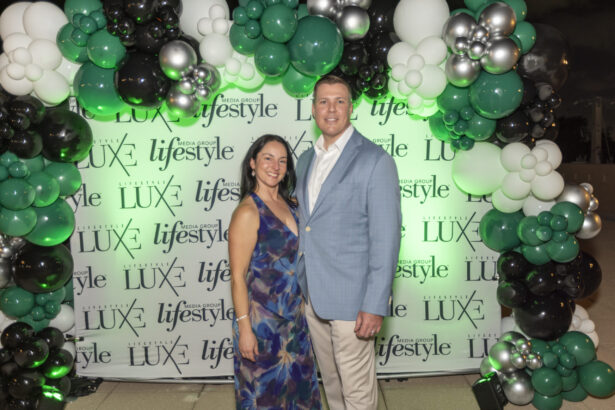 A smiling couple poses in front of a step-and-repeat banner with "LUXE" and "lifestyle" logos, surrounded by an arch of green, black, white, and silver balloons at the Influential People 2025 outdoor event. Lifestyle