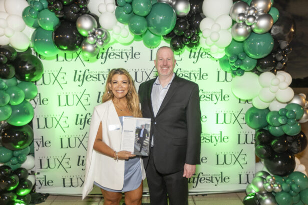A smiling woman holding a magazine stands next to a smiling man in a suit, both posing as part of the 2025 People of Influence event in front of a green, white, and black balloon arch and a "Lifestyle Media Group LUX" backdrop. Lifestyle