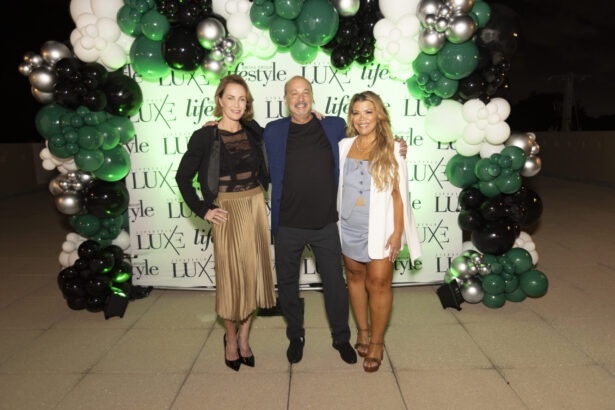 Three people smile and pose in front of a green and white balloon arch and a photo backdrop featuring "LUX," "lifestyle," and the 2025 Women of Influence celebration at an outdoor event held at night. Lifestyle