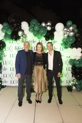 Three people dressed in semi-formal attire stand smiling in front of a green, white, and black balloon arch and a "LUXE Lifestyle" backdrop at the 2025 Women of Influence outdoor event held at night. Lifestyle