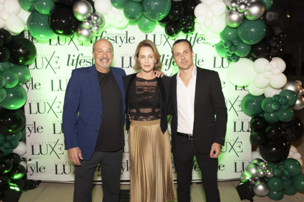 Three people stand smiling in front of a backdrop with "Lifestyle LUXE" logos and 2025 Men of Influence and Women of Influence signage, surrounded by green, black, silver, and white balloon decorations at an event. Lifestyle
