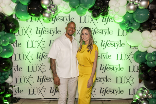 A man in a white outfit and a woman in a yellow dress, honorees of the Men of Influence and Women of Influence 2025, stand smiling under a green, black, and white balloon arch beside a step-and-repeat with "Lifestyle" and "LUXE" logos. Lifestyle