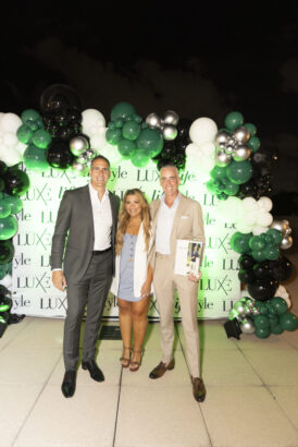 Three influential people dressed in business attire stand and smile in front of a "LUXE" backdrop with green, white, and black balloons at an outdoor event celebrating the 2025 Men of Influence and 2025 Women of Influence. Lifestyle