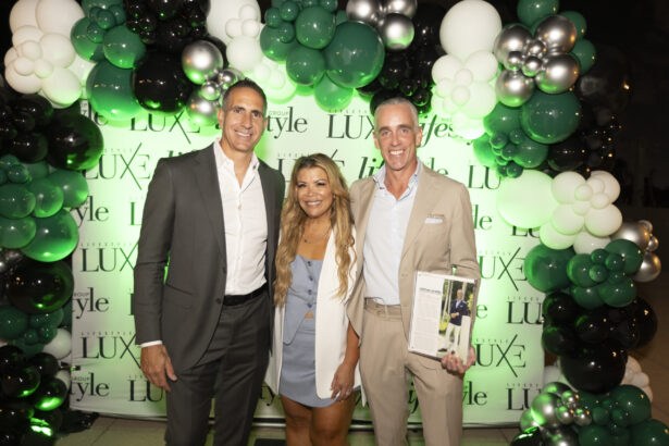 Three people in business attire smile before a green, white, and black balloon arch with a “LUXE” and “Style” backdrop. One proudly holds a magazine featuring a 2025 Men of Influence cover. Lifestyle