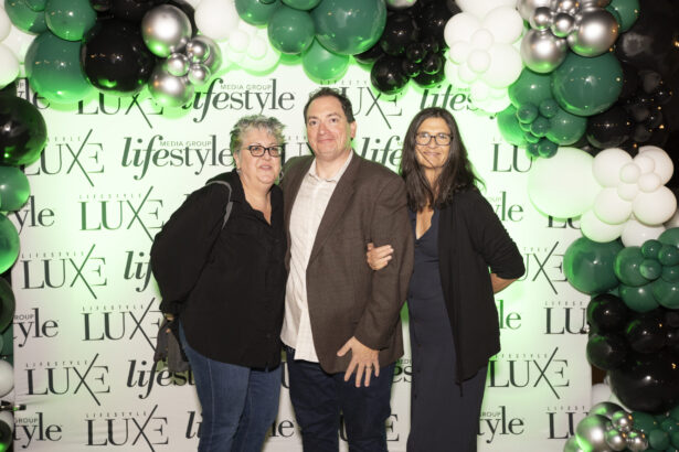 Three people pose together, smiling, in front of a step-and-repeat backdrop with "LUXE," "lifestyle," and 2025 Men of Influence logos. Black, white, and green balloons frame the top and sides of the backdrop. Lifestyle