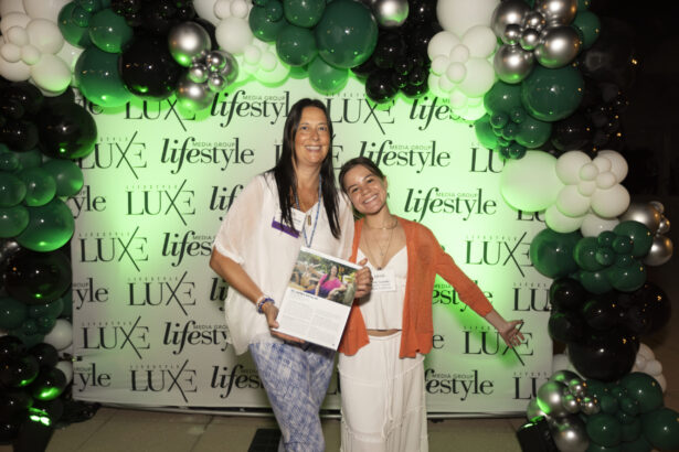 Two women smile and pose in front of a black, white, and green balloon arch and a "LUX lifestyle" step-and-repeat. One holds an open magazine as they celebrate the 2025 Women of Influence event, both looking happy and inspired. Lifestyle