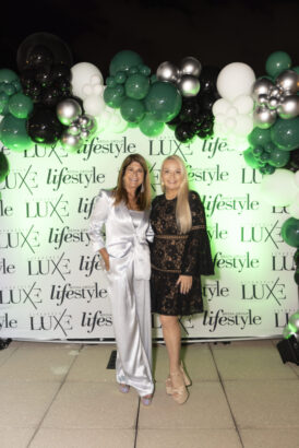 Two women, candidates for 2025 Women of Influence, stand smiling in front of a "LUXE lifestyle" logo backdrop adorned with green, black, and white balloon clusters. One wears a silver suit; the other stuns in a black lace dress. Lifestyle