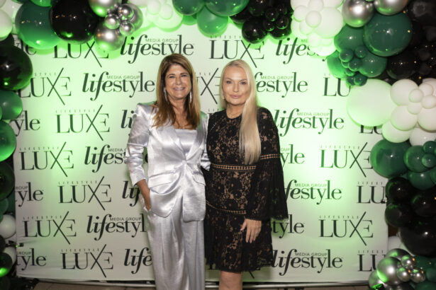 Two women stand smiling in front of a step-and-repeat backdrop with "LUXE" and "lifestyle" logos, surrounded by black, white, and green balloons at the 2025 Women of Influence event. Lifestyle