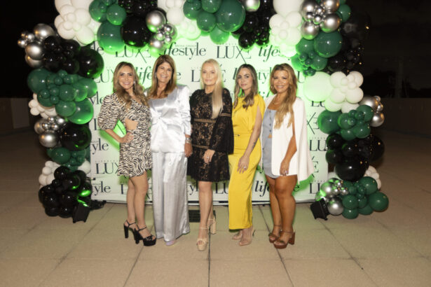 Five women stand smiling in front of a green and white balloon arch and a "LUX lifestyle" step-and-repeat backdrop at the 2025 Women of Influence outdoor evening event, dressed in stylish, semi-formal outfits. Lifestyle