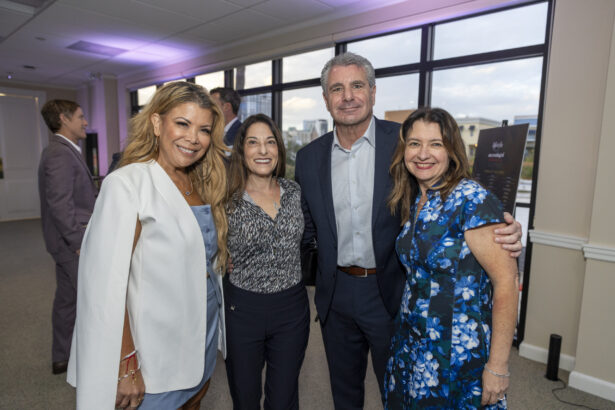 Four adults, three women and one man, stand together indoors at a Women of Influence 2025 event, smiling at the camera. They are dressed in business or semi-formal attire, with large windows and evening light in the background. Lifestyle