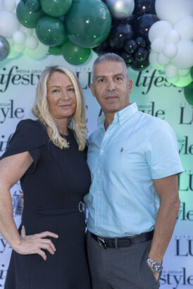 A woman in a black dress and a man in a light blue shirt pose together in front of a "Lifestyle" backdrop with green, black, and white balloons at the 2025 Women of Influence event. Lifestyle