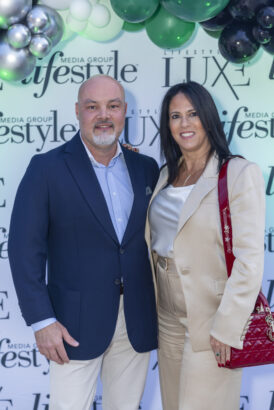 A man in a navy blazer and beige pants stands next to a woman in a beige suit with a red handbag, posing at the 2025 "Lifestyle Luxe Media Group" step-and-repeat, celebrating Men of Influence and Women of Influence beneath green and silver balloons. Lifestyle