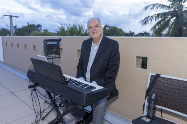 A smiling man in a blazer sits at an electric keyboard on an outdoor patio, with a Men of Influence 2025 sign beside his speaker and tablet. Palm trees and a cloudy sky create a lively backdrop. Lifestyle
