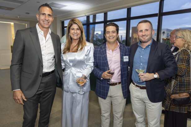 Four people, including honorees from the Women of Influence and Men of Influence 2025 lists, pose together at an indoor event in business attire, holding drinks and smiling at the camera. Other guests and large windows appear in the background. Lifestyle